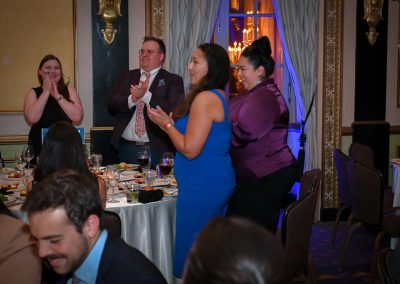 The image shows a group of people gathered in a room. They are attending the MALDEF Chicago Gala in 2024. The setting includes a table with food and tableware, and the people are smiling. The individuals are dressed in party attire, indicating a festive occasion.