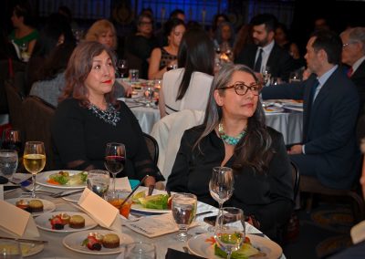 The image depicts women sitting at a table during the MALDEF Chicago Gala 2024. The table is set with food, drinks, and tableware, while people are seen enjoying a meal at a restaurant event.