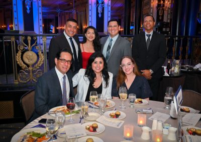 The photo is of a group of people posing for a photo. They are indoors, at a banquet or rehearsal dinner, standing around a dining table with wine glasses and champagne stemware. The people in the photo are dressed in formal clothing and are smiling.