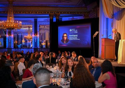 The image shows a person giving a presentation at the MALDEF OGALI 9024 Chicago Awards. The individual receiving the award is Rodriguez, a singer and songwriter. The event appears to be a function hall with an audience present.