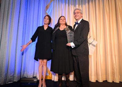 The image depicts a group of people posing for a photo at the MALDEF Achievement event in Chicago, Illinois on June 13, 2024. The individuals are dressed in formal attire and are standing in front of a theater curtain. The photo captures the people smiling and dressed elegantly. Tags include clothing, person, suit, smile, dress, man, woman, indoor, human face, formal wear, theater curtain, footwear, standing, curtain, wall, and people.